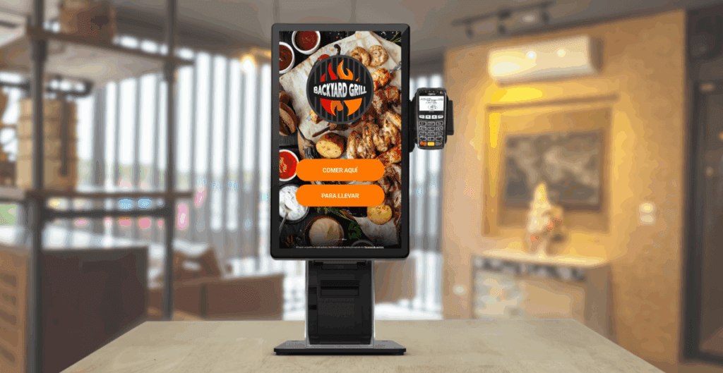 A self-ordering kiosk at a modern grill restaurant with a Backyard Grill interface and a card reader attached, positioned on a wooden table.